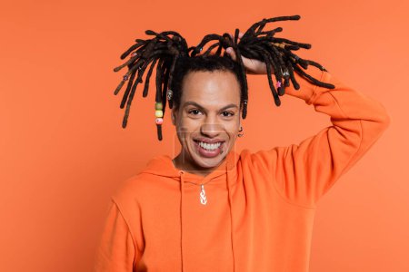 happy multiracial man touching dreadlocks on coral background