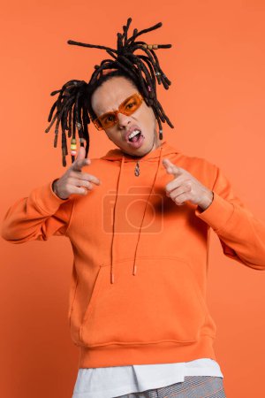 pierced multiracial man in hoodie wearing stylish sunglasses and pointing with fingers isolated on coral background