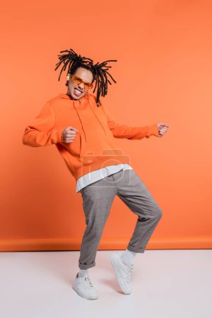 excited multiracial man in hoodie wearing stylish sunglasses and dancing on coral background
