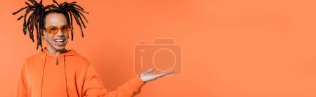 happy multiracial man in hoodie and stylish sunglasses pointing with hand on coral background, banner 