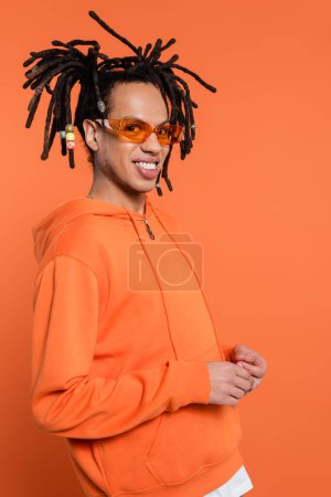 joyful multiracial man in hoodie and stylish sunglasses smiling isolated on coral 