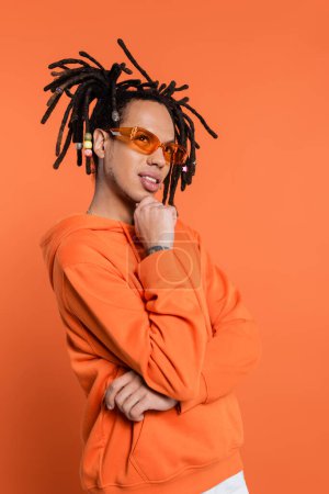 Téléchargez les photos : Thoughtful multiracial man in hoodie and stylish sunglasses on coral background - en image libre de droit