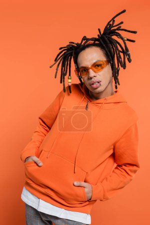 pierced multiracial man in hoodie and stylish sunglasses posing with hands in pockets on coral background