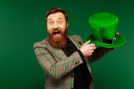 Excited bearded man holding hat while celebrating saint patrick day isolated on green 
