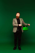 Full length of bearded man in jacket holding hat during saint patrick day on green background  tote bag #640344874