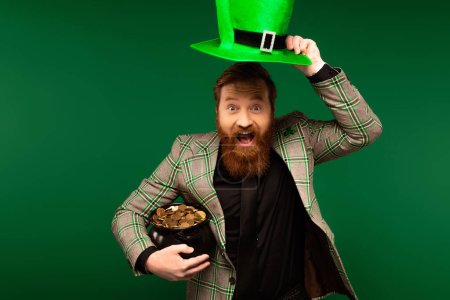 Excited bearded holding hat and pot with coins during saint patrick day isolated on green 