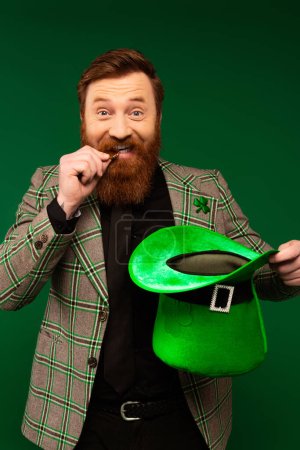 Bearded man biting coin and holding hat during saint patrick day isolated on green 