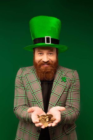 Foto de Smiling bearded man in hat holding coins while celebrating saint patrick day isolated on green - Imagen libre de derechos