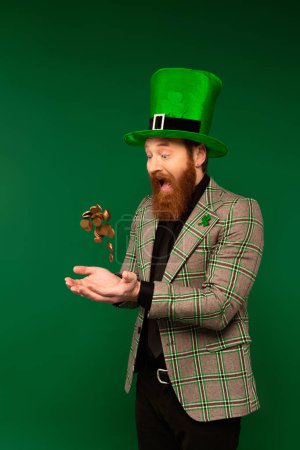 Téléchargez les photos : Choqué homme barbu dans le chapeau jetant des pièces pendant saint Patrick jour isolé sur vert - en image libre de droit