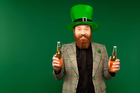 Foto de Smiling bearded man in hat holding bottles of beer isolated on green - Imagen libre de derechos