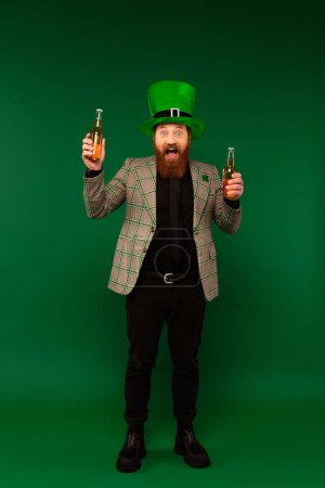 Photo for Full length of astonished man in hat holding bottles of beer on green background - Royalty Free Image