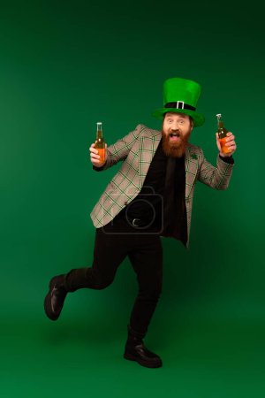 Foto de Longitud completa del hombre excitado en sombrero celebrando el día de San Patricio y sosteniendo botellas de cerveza sobre fondo verde - Imagen libre de derechos