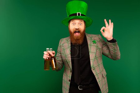 Foto de Excited man in hat holding bottles of beer and showing okay gesture isolated on green - Imagen libre de derechos