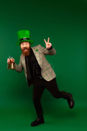 Excited bearded man in hat holding bottles of beer and showing peace sign on green background 