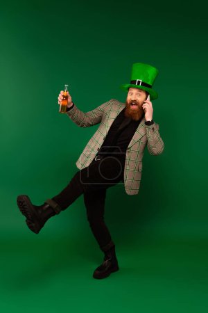 Foto de Excited bearded man in hat holding beer and talking on smartphone on green background - Imagen libre de derechos