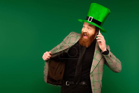Foto de Excited bearded man in jacket talking on smartphone during saint patrick day isolated on green - Imagen libre de derechos