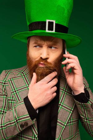 Téléchargez les photos : Thoughtful bearded man in hat with clover talking on smartphone isolated on green - en image libre de droit
