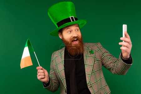 Sonriente hombre barbudo con sombrero sosteniendo la bandera irlandesa y tomando selfie en el teléfono inteligente aislado en verde 