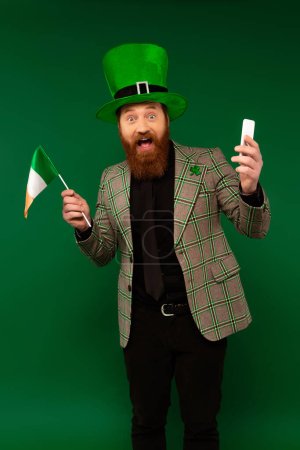 Excited man in hat with clover holding smartphone and Irish flag isolated on green 