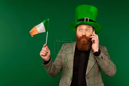 Hombre barbudo con sombrero hablando en smartphone y mirando la bandera irlandesa aislada en verde 