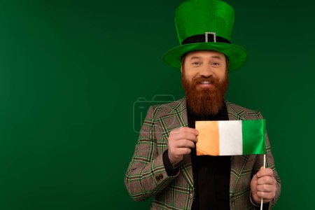 Hombre positivo con sombrero sosteniendo bandera irlandesa durante la celebración del día de San Patricio aislado en verde 