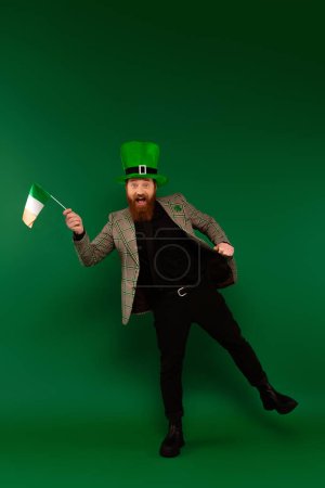 Longitud completa de hombre barbudo alegre en sombrero con bandera irlandesa sobre fondo verde 