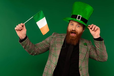 Excited bearded man in hat holding Irish flag during saint patrick day isolated on green 