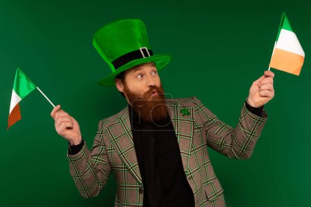Excited bearded man in hat holding Irish flags isolated on green 