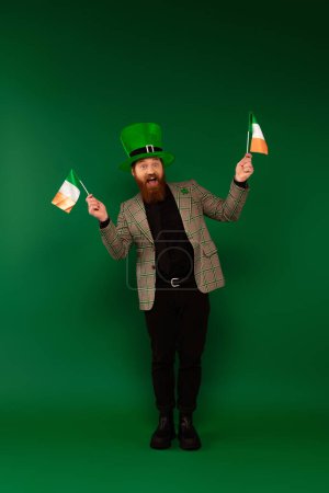 Longitud completa de hombre barbudo alegre con sombrero sosteniendo banderas irlandesas sobre fondo verde 