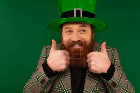 Téléchargez les photos : Smiling bearded man in hat showing like gesture isolated on green - en image libre de droit
