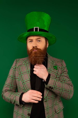 Focused man in jacket with clover and hat touching tie isolated on green 