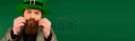 Téléchargez les photos : Portrait of man in hat touching moustache and looking at camera isolated on green, banner - en image libre de droit