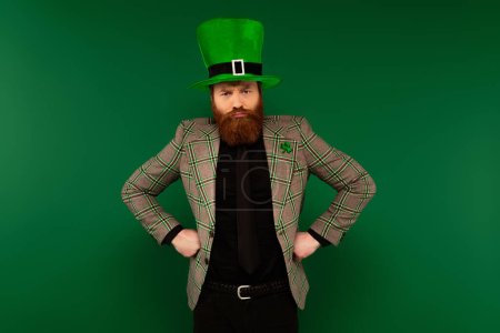 Angry bearded man in hat with clover holding hands on hips isolated on green 