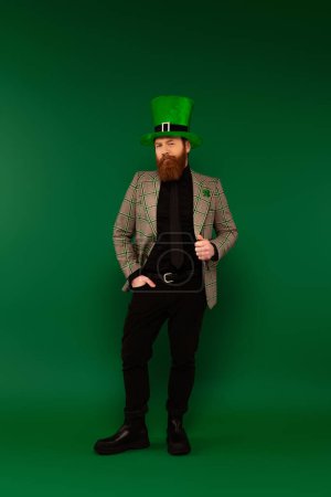Full length of bearded man in clover on jacket and on hat posing on green background 