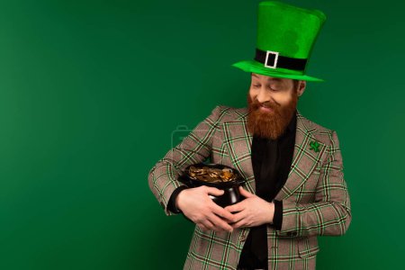 Smiling bearded man in hat holding pot with coins isolated on green 