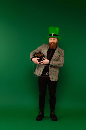 Cheerful bearded man in hat holding pot with golden coins on green background 