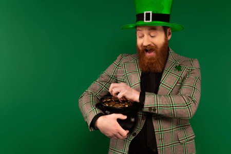 Téléchargez les photos : Homme excité dans le chapeau tenant pot avec des pièces d'or pendant saint Patrick jour isolé sur vert - en image libre de droit