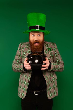 Bearded man in hat with clover holding pot with golden coins isolated on green 