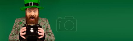Photo for Astonished man in hat looking at pot with golden coins isolated on green, banner - Royalty Free Image