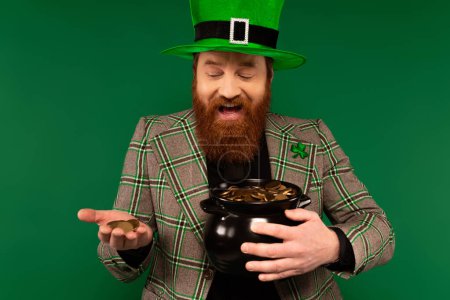Cheerful man in hat holding golden coins and pot during saint patrick day isolated on green 