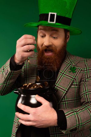Foto de Excited man in hat with clover pouring coins in pot isolated on green - Imagen libre de derechos