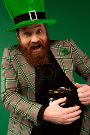 Foto de Amazed man in hat with clover holding pot with golden coins isolated on green - Imagen libre de derechos