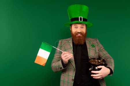 Hombre positivo en sombrero con trébol sosteniendo bandera irlandesa y olla con monedas aisladas en verde 