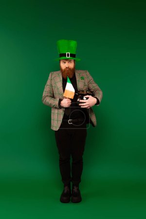 Pensive man in hat holding Irish flag and pot with coins on green background 