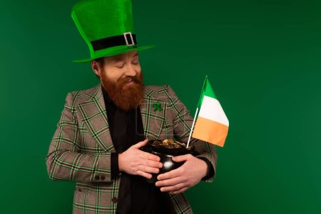 Positive Mann mit Hut hält Topf mit Münzen und irische Flagge isoliert auf grün 