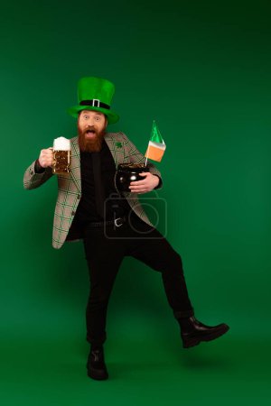 Hombre excitado en sombrero sosteniendo cerveza y olla con monedas y bandera irlandesa sobre fondo verde 