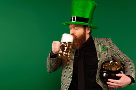 Hombre barbudo en sombrero bebiendo cerveza y sosteniendo la olla con monedas aisladas en verde 