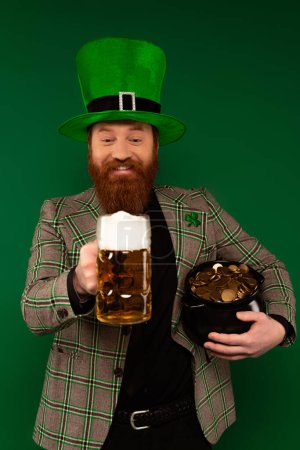 Fröhlicher bärtiger Mann mit Hut, der ein Glas Bier und einen Topf mit Münzen auf Grün hält 
