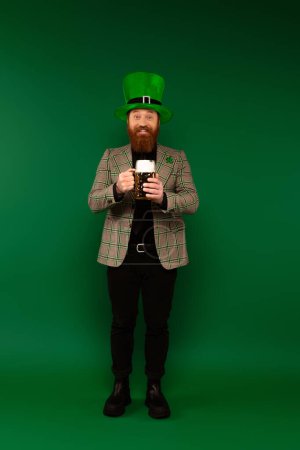 Voller Länge lächelnder bärtiger Mann mit Hut, der ein Glas Bier auf grünem Hintergrund hält 