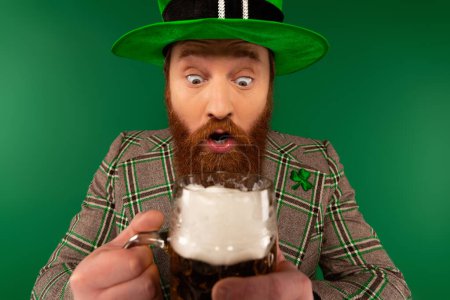 Schockierter Mann mit Hut und Kleeblatt auf Jacke hält verschwommenes Bierglas auf Grün 